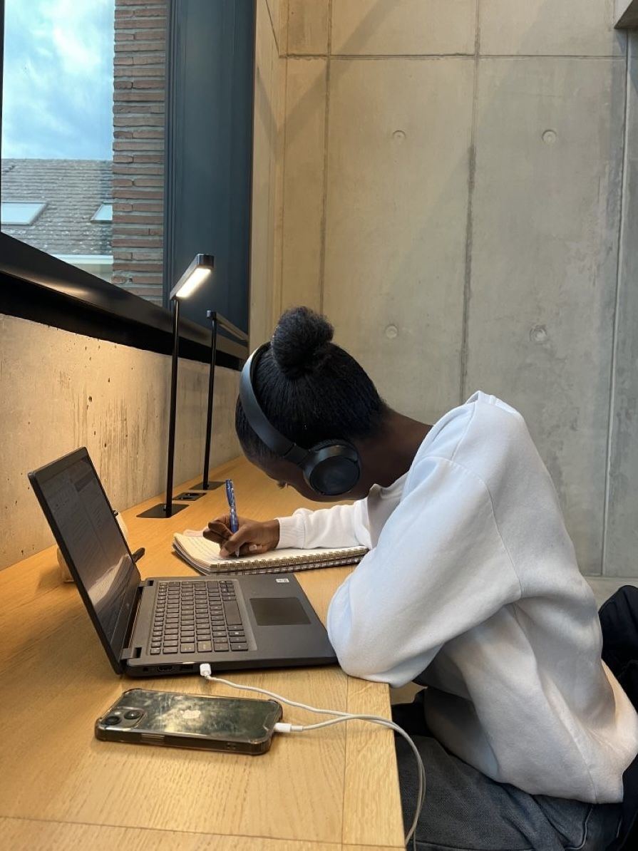 A woman in headphones writing in a notebook beside her laptop at a sunlit desk