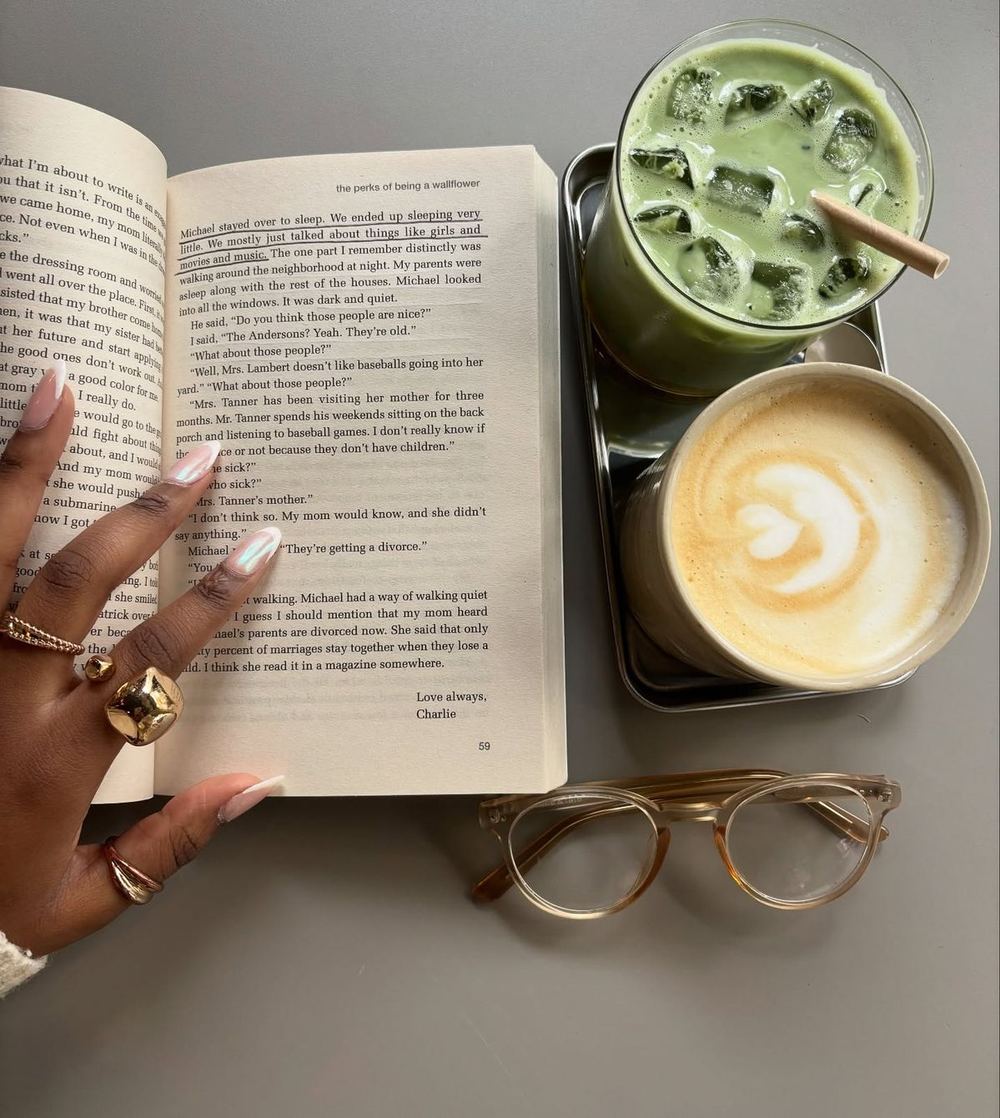 A hand with gold rings resting on an open book beside an iced matcha and a latte